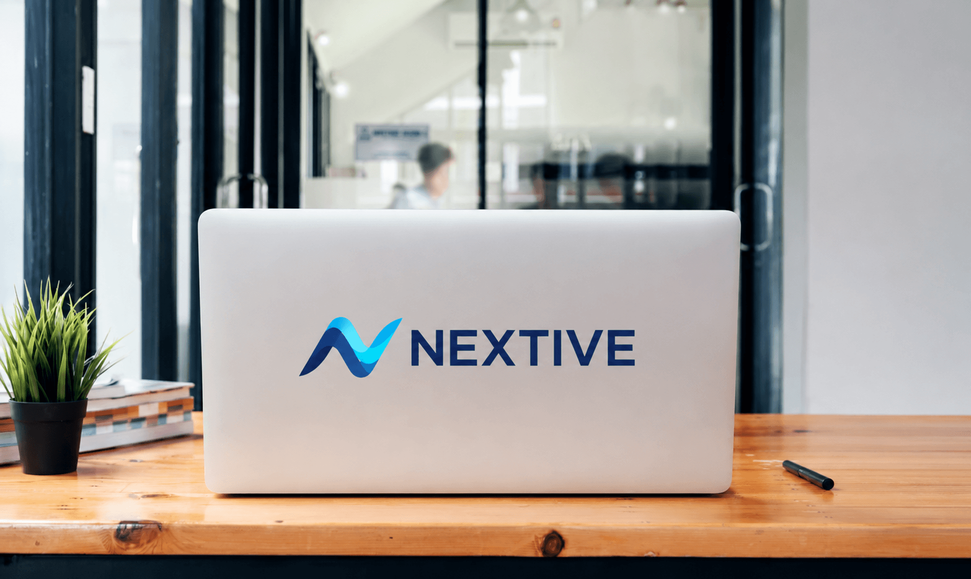 NEXTIVEのロゴが入ったノートパソコンがオフィスのデスクに置かれ、奥で社員たちが働いている様子